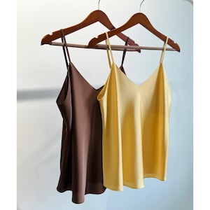 Puede incluir: Dos camisetas sin mangas colgadas en perchas de madera. Una es de color marrón liso y la otra de color amarillo liso. Ambas camisetas tienen un diseño de cuello en V y tirantes finos. Las camisetas están hechas de un material sedoso.