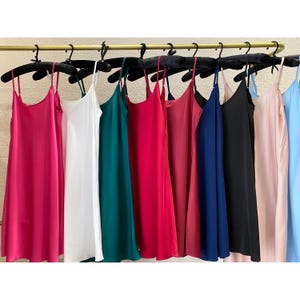 Pode incluir: Uma coleção de vestidos tipo slip sem mangas pendurados em cabides pretos numa barra dourada. Os vestidos estão disponíveis numa variedade de cores sólidas, incluindo magenta, branco, verde-azulado, vermelho, rosa escuro, azul-marinho, preto, rosa claro e azul claro.