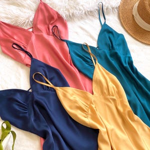 Pode incluir: Uma coleção de vestidos de seda em tons de coral, verde-azulado, azul marinho e dourado. Os vestidos têm alças finas e uma silhueta fluida. Um chapéu de sol de palha com uma faixa branca também é visível no fundo.