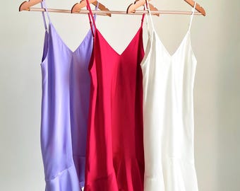 May include: Three sleeveless slip dresses hanging on wooden hangers. The dresses are in solid lavender, bright magenta, and white. Each dress features a V-neckline, thin adjustable spaghetti straps, and a ruffled hemline at the bottom edge.
