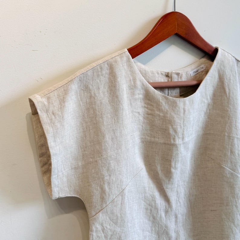 May include: A light beige linen top with short sleeves, hanging on a wooden hanger. The top has a round neckline and a subtle texture. The garment is simple and elegant, suitable for casual wear.