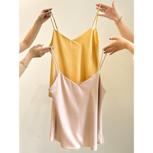 Puede incluir: Dos tops de seda tipo camisola. Uno es rosa pálido, el otro amarillo dorado. Ambos tienen cuello en V y finas correas de espagueti. Las manos sostienen las prendas, mostrando la tela y el diseño.