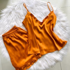 May include: A set of orange satin pajamas, including a camisole top with thin straps and ruffled trim, and matching shorts. The sleepwear is displayed on a white, fluffy surface.