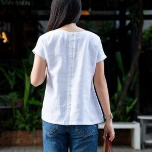 May include: A white short-sleeved top featuring a vertical row of small matching fabric-covered buttons running down the center of the back. The garment has a round neckline and a relaxed fit, paired with blue denim jeans and a woven straw bag.