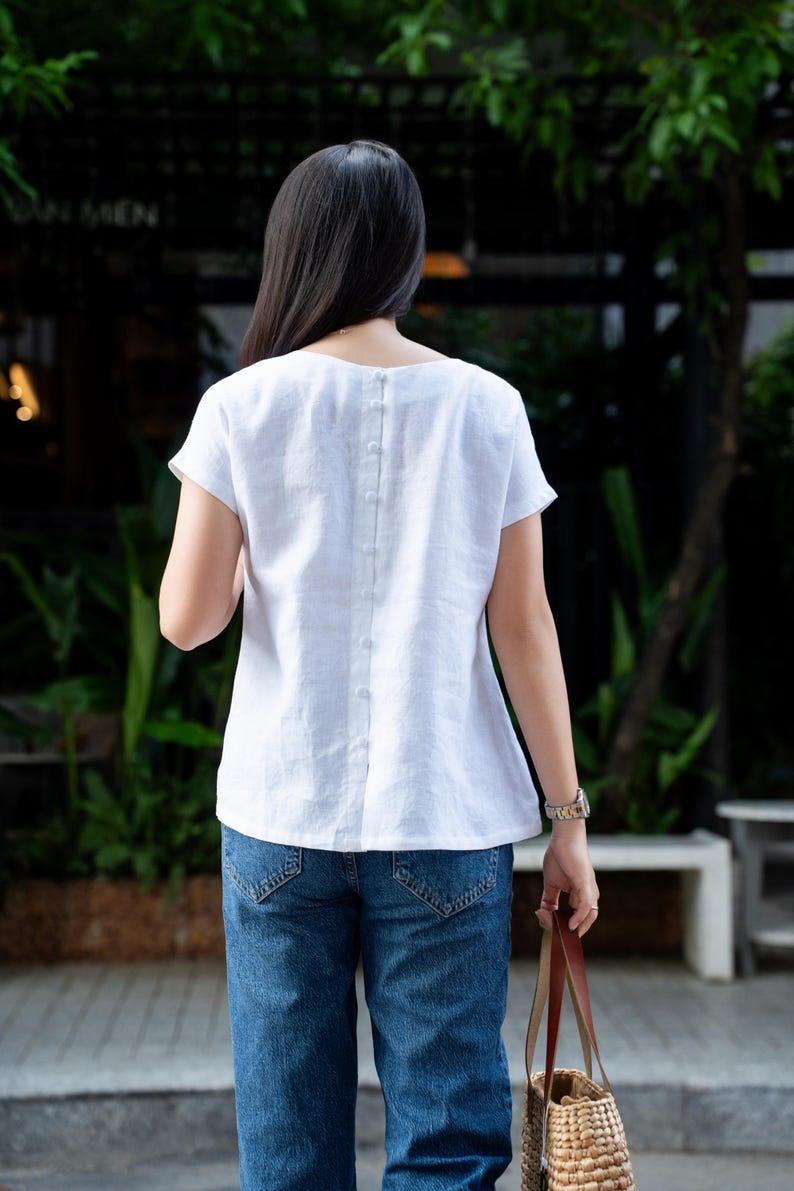 May include: A white short-sleeved top featuring a vertical row of small matching fabric-covered buttons running down the center of the back. The garment has a round neckline and a relaxed fit, paired with blue denim jeans and a woven straw bag.