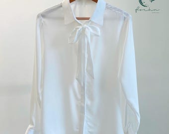 May include: A white long-sleeve blouse with a tie-neck detail, hanging on a wooden hanger. The blouse has a classic collar and buttoned cuffs. The fabric appears to be a smooth, lightweight material.