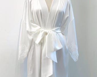Könnte beinhalten: Ein weißer Satin-Bademantel mit langen Ärmeln, Spitzenbesatz an den Manschetten und einem passenden Spitzenrand am Saum. Der Bademantel verfügt über einen breiten Satin-Bindegürtel in der Taille, einen V-Ausschnitt und eine glatte, glänzende Oberfläche.