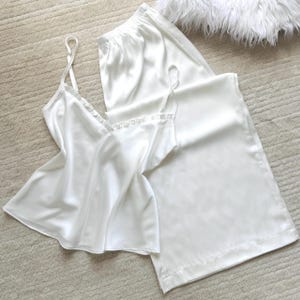 May include: A white satin pajama set, consisting of a camisole top with lace trim and spaghetti straps, and matching wide-leg pants. The set is displayed on a neutral-colored surface, with a fluffy white accent in the background.