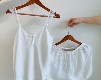 May include: A white satin pajama set, including a camisole top with lace trim and a small bow, and matching shorts. The set is displayed on wooden hangers against a plain background.