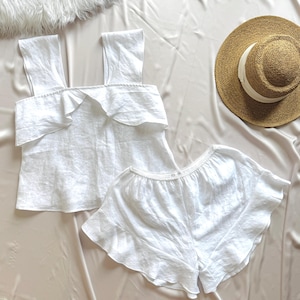 Puede incluir: Un conjunto de dos piezas blanco que consta de una blusa sin mangas con tirantes anchos y un volante sobre el pecho, combinado con pantalones cortos blancos a juego con dobladillo con volantes, junto a un sombrero de paja beige con cinta blanca.