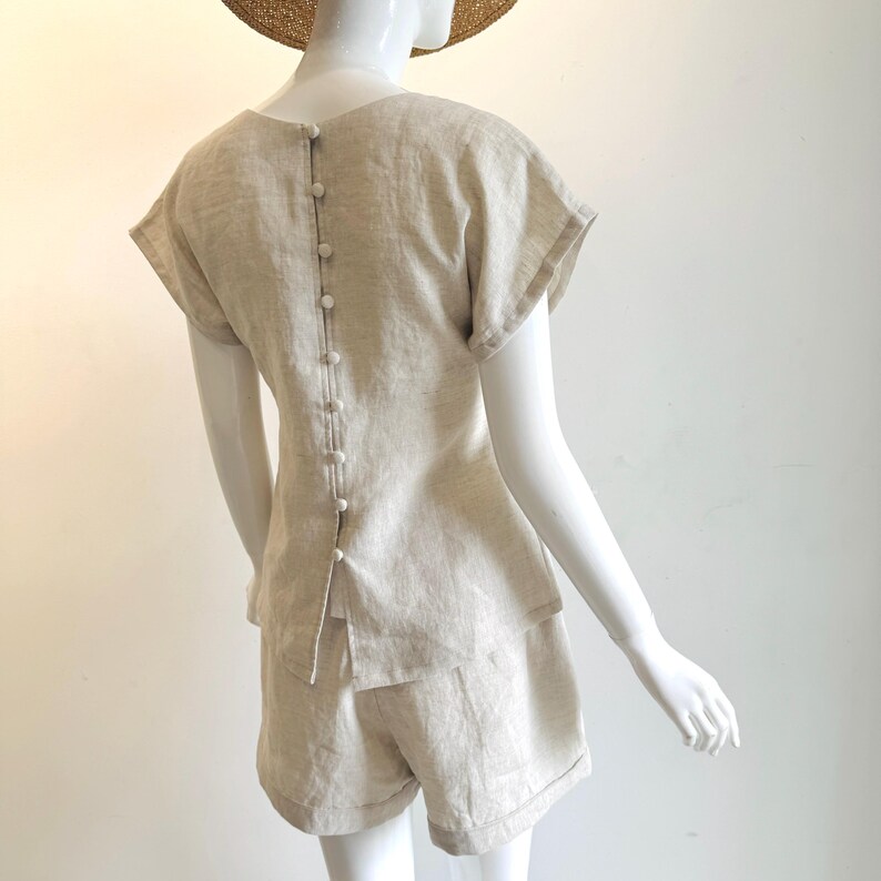 May include: A beige linen shorts set, comprising a short-sleeved top with buttons down the back and matching shorts. The outfit is displayed on a mannequin. The fabric appears lightweight, suitable for warmer weather. The set is made of linen.