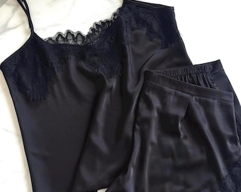 May include: A black two-piece pajama set consisting of a camisole and shorts. The camisole features thin adjustable straps and black lace trim along the neckline. The matching shorts have an elastic waistband and black lace trim on the hem.
