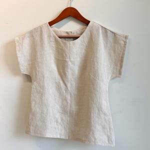May include: A light beige linen top with short sleeves, hanging on a wooden hanger. The top has a round neckline and a simple, minimalist design. The fabric appears to be lightweight and breathable, suitable for warm weather.