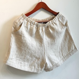 May include: Natural linen shorts with an elasticated waistband and side pockets, displayed on a wooden hanger. These shorts have a relaxed fit and a simple design, suitable for casual wear.