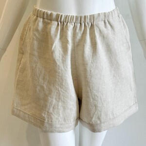 May include: Beige linen shorts with an elasticated waistband. The shorts have a relaxed fit and a simple design, suitable for casual wear. The fabric appears to be lightweight and breathable, ideal for warm weather. The shorts are displayed on a mannequin.