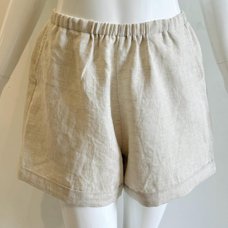 May include: Beige linen shorts with an elasticated waistband. The shorts have a relaxed fit and a simple design, suitable for casual wear. The fabric appears to be lightweight and breathable, ideal for warm weather. The shorts are displayed on a mannequin.