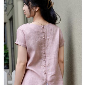 May include: A short-sleeved, dusty pink blouse featuring a vertical row of matching fabric-covered buttons running down the center of the back. The garment has a round neckline, a straight hemline with a small back slit, and a textured fabric finish.