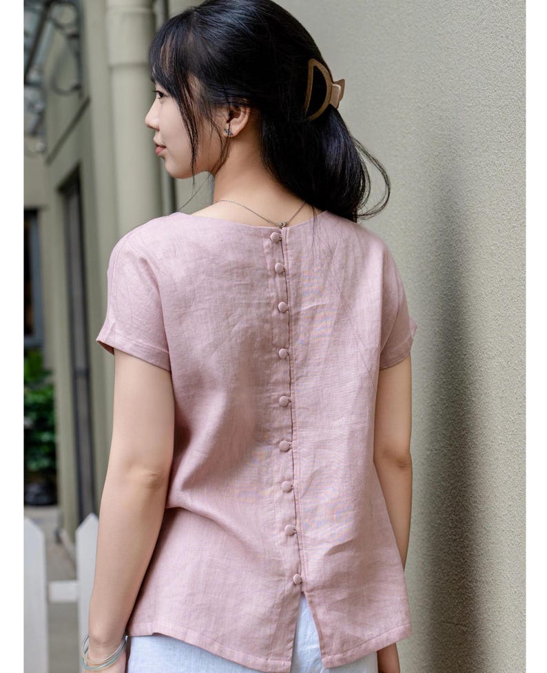 May include: A short-sleeved, dusty pink blouse featuring a vertical row of matching fabric-covered buttons running down the center of the back. The garment has a round neckline, a straight hemline with a small back slit, and a textured fabric finish.