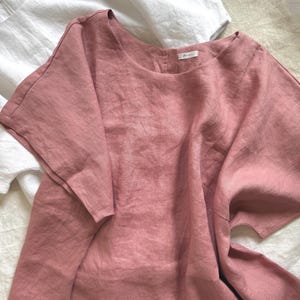 May include: A dusty rose-colored linen top with short, wide sleeves and a round neckline. The garment features a button-down back closure and a small white brand label at the inner neck. A white linen top is partially visible underneath the rose top.