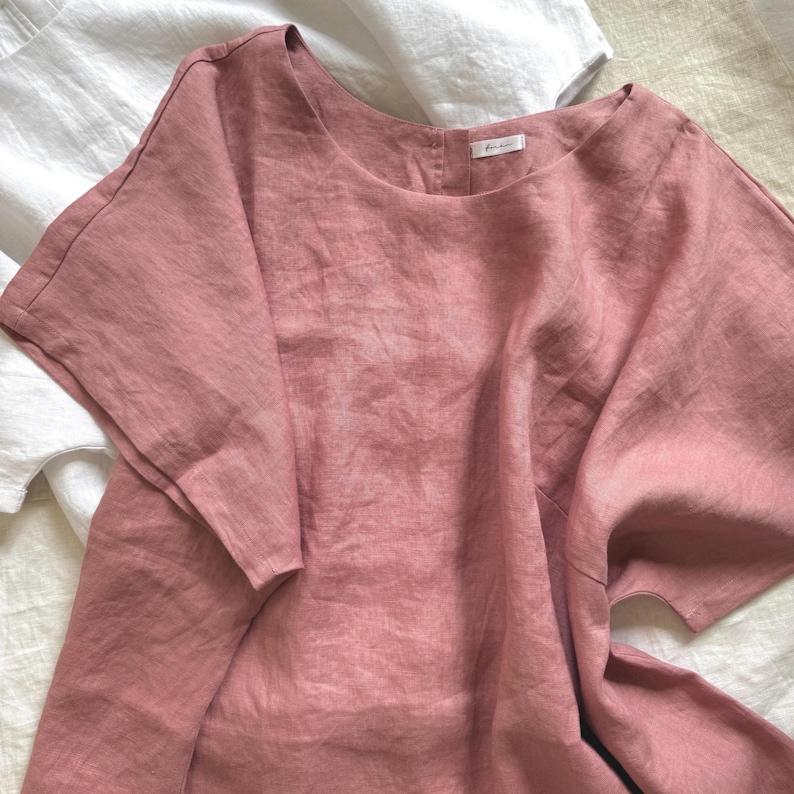 May include: A dusty rose-colored linen top with short, wide sleeves and a round neckline. The garment features a button-down back closure and a small white brand label at the inner neck. A white linen top is partially visible underneath the rose top.