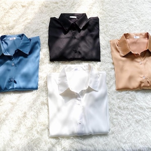 Puede incluir: Cuatro camisas de manga larga con botones en azul, negro, blanco y beige. Cada camisa presenta un cuello clásico, una tapeta de botones frontal y un acabado suave similar al satén. Las camisas están dispuestas en un grupo ordenado.