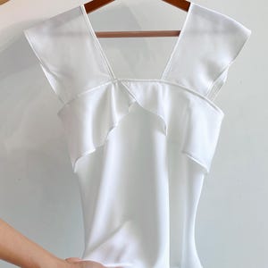 May include: A white sleeveless top with a square neckline and ruffled detailing. The top is made of a smooth, flowing fabric and hangs on a wooden hanger.
