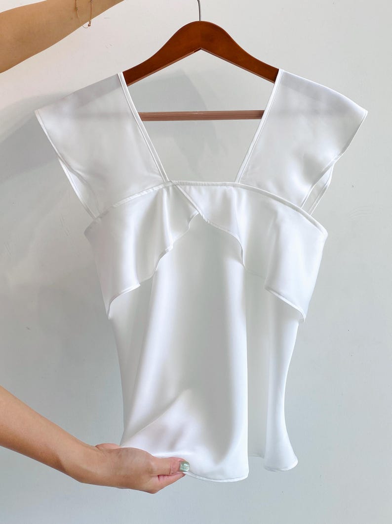 May include: A white sleeveless top with a square neckline and ruffled detailing. The top is made of a smooth, flowing fabric and hangs on a wooden hanger.