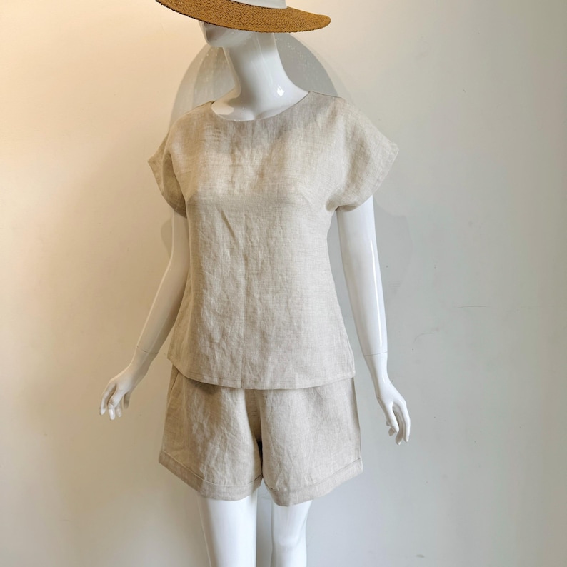 May include: A beige linen shorts set on a mannequin. The outfit comprises a short-sleeved top and matching shorts. A straw hat with a white band is on the mannequin's head. The set is made from a lightweight, breathable fabric, ideal for warm weather.