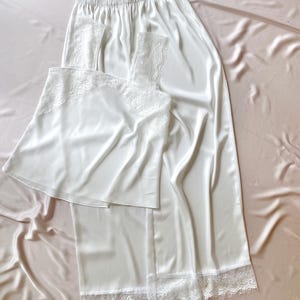 May include: A white satin pyjama set, comprising a camisole and trousers, adorned with delicate lace trim. The set is presented on a light pink satin surface, creating a soft and elegant aesthetic. The camisole and trousers are made of a smooth, flowing fabric.