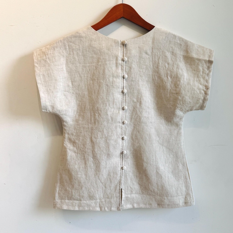 May include: A light beige linen top with short sleeves and a row of small buttons down the back. The top is hanging on a wooden hanger. The fabric has a textured appearance.