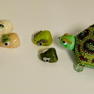 May include: A collection of decorative turtle figurines. The larger turtle has a beaded shell in shades of green, yellow, and brown. Smaller turtle-shaped stones in various colors are also present, all with painted eyes and green accents.