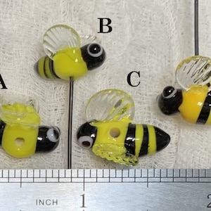 May include: Four glass bee beads with black and yellow stripes and clear wings. The beads are approximately 1 inch in length. The beads are on a white surface with a ruler for scale.