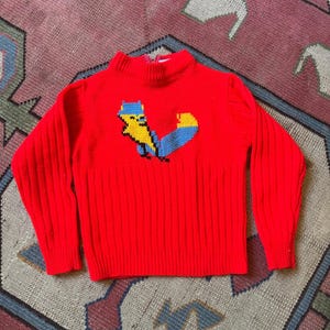 May include: A bright red long-sleeve sweater featuring a ribbed texture and a mock neck collar. The center of the chest displays a knitted graphic of a yellow and blue creature with a large tail. The garment has a zipper closure at the back neck.