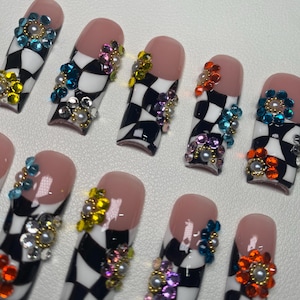 May include: Set of ten long, square-shaped false nails with a black and white checkered pattern. Each nail is decorated with colorful rhinestones and pearls.