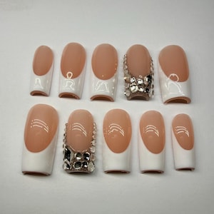 May include: A set of ten artificial nails with a French manicure design. The nails have a nude base with white tips. Two nails are embellished with rhinestones. The nails are oval-shaped.