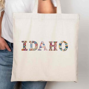 Puede incluir: Bolso tote de color crema con la palabra "IDAHO" en letras coloridas y estampadas. El bolso tiene asas largas. Las letras están rellenas de diseños florales y geométricos. El bolso lo sostiene una persona.