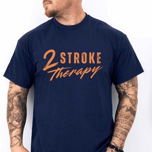 Może przedstawiać: Koszulka w kolorze granatowym z napisem "2 STROKE therapy" w kolorze pomarańczowym.