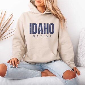 May include: A beige hoodie with the text "IDAHO NATIVE" in navy blue. The hoodie has a front pocket and a hood. The person in the image is wearing light blue jeans with distressed details.