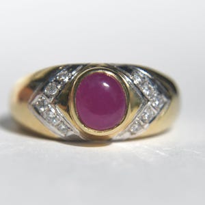 May include: A gold ring featuring a central oval cabochon-cut deep pink gemstone set in a yellow gold bezel. The band is accented with two V-shaped channels of small clear stones set in white metal, contrasting with the polished yellow gold shank.