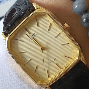 May include: A gold-tone Citizen Forma watch with a rectangular case and a textured champagne-colored dial featuring thin gold hour markers. The watch has a black leather strap with a crocodile-embossed pattern and a small gold crown on the side.