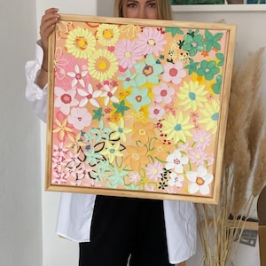 Könnte beinhalten: Quadratische Blumenkunst in einem Holzrahmen. Das Kunstwerk zeigt eine Vielzahl farbenfroher Blumen in Rosa-, Gelb-, Grün- und Weißtönen vor einem pfirsichfarbenen Hintergrund. Der Rahmen hat eine hellbraune Farbe.