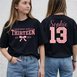 May include: Black t-shirt with pink text and graphics. The front of the shirt reads "BIRTHDAY GIRL THIRTEEN EST. 2013" with a bow graphic. The back of the shirt has the name "Sophie" and the number "13" in a pink leopard print.