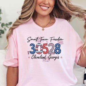 May include: A light pink t-shirt with the text "Small Town Freedom" and the zip code "30528" in red, white, and blue. The text "Cleveland, Georgia" is below the zip code, with fireworks and star accents.