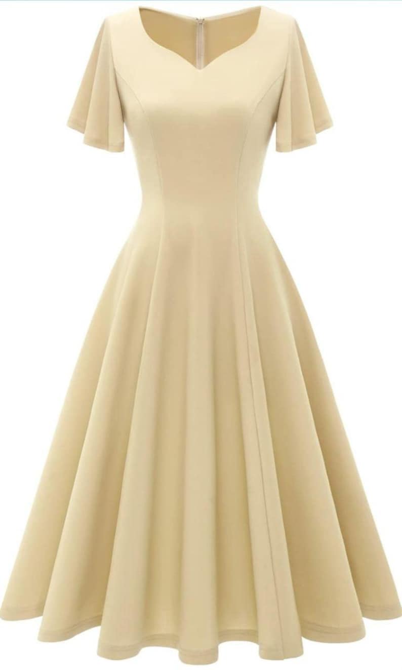 Puede incluir: Un vestido beige claro con escote de coraz&oacute;n y mangas cortas acampanadas. El vestido tiene un corpi&ntilde;o ajustado y una falda fluida.