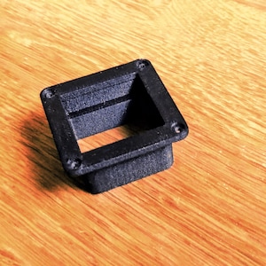Könnte beinhalten: Ein schwarzes, quadratisches 3D-gedrucktes Bauteil mit hohlem Zentrum und vier kleinen Löchern in den Ecken. Das Bauteil liegt auf einer Holzoberfläche.