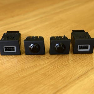 Könnte beinhalten: Vier schwarze Kfz-Schalter sind auf einer Holzoberfläche angeordnet. Zwei haben einen USB-Anschluss, und zwei haben einen runden Knopf. Die Schalter sind rechteckig mit einer dunkelgrauen Front und einem schwarzen Gehäuse.