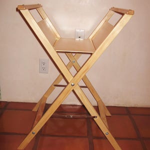 May include: A light-colored wooden folding easel with a rectangular shelf. The easel has an X-frame design with a horizontal bar for stability. The easel is made of light-colored wood and has a natural finish. The easel is designed to hold a canvas or artwork.