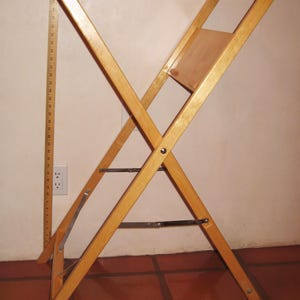 May include: A wooden folding stand, approximately 36 inches tall, with an X-frame design. The stand is made of light-colored wood and features a small wooden panel. Metal bars provide structural support.