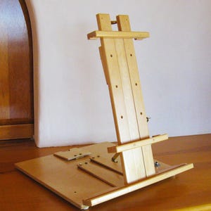 May include: A wooden artist's easel, constructed from light-colored wood. The easel features an adjustable angle and a base for supporting a canvas. The design includes a rectangular base and a vertical support structure.