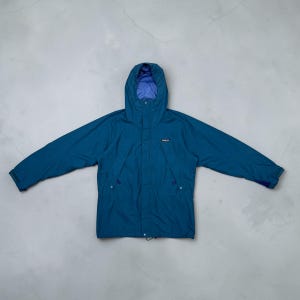 Könnte beinhalten: Eine petrolfarbene Kapuzenjacke mit Frontreißverschluss, hohem Kragen und violettem Innenfutter. Das Kleidungsstück verfügt über lange Ärmel mit verstellbaren Bündchen und einen kleinen, rechteckigen Logo-Aufnäher auf der Brust.